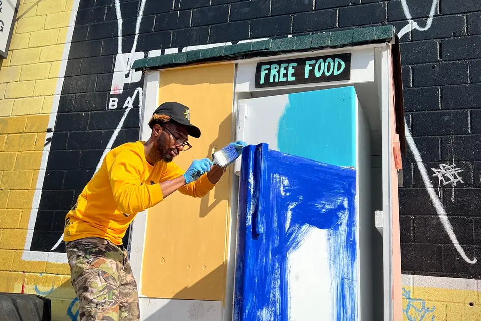 A man in working clothes stands in the hot sun painting the doors of a fridge blue. The a sign with the words "Free Food" hangs on the lintel of the fridge shed.
