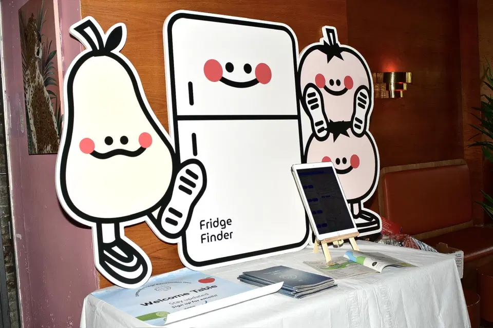 A table on top of which stands a cardboard cutout of the Fridge Finder mascots. On top of the table are printed booklets, a welcome poster, and an iPad on a display stand.
